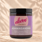 Eleven TLC Organic Ashwagandha Root Powder 120g amber glass jar with a black lid on travertine stone against a warm cream plaster wall, with a soft shadow to the right.