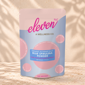 Eleven TLC Blue Spirulina Powder 500g light blue and pink pouch standing on a travertine ledge against warm plaster, with a contact shadow beneath the base and a soft shadow to the right.