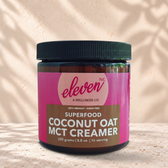Eleven TLC Coconut Oat MCT Creamer 200g amber jar on beige travertine against warm plaster, with a dark base shadow and soft rightward shadow.