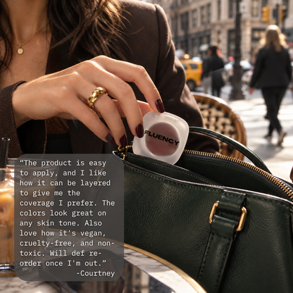 woman's hand with gold ring and dark nail polish pulling out a fluency blush palette from her black purse sitting at a cafe with coffee on a busy street in nyc