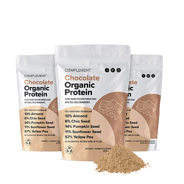 Three bags of Complement Chocolate Organic Protein powder are displayed, featuring plant-based ingredients like almond, chia seed, flax seed, pumpkin seed, sunflower seed, and yellow pea. A small pile of the powder is in front.