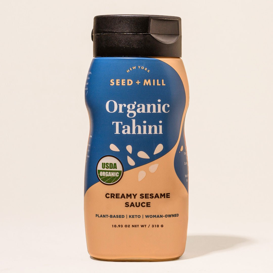 A bottle of Seed + Mill Organic Tahini with a blue label, displaying the product name and the description 'Creamy Sesame Sauce'. The bottle is upright and has a black cap.