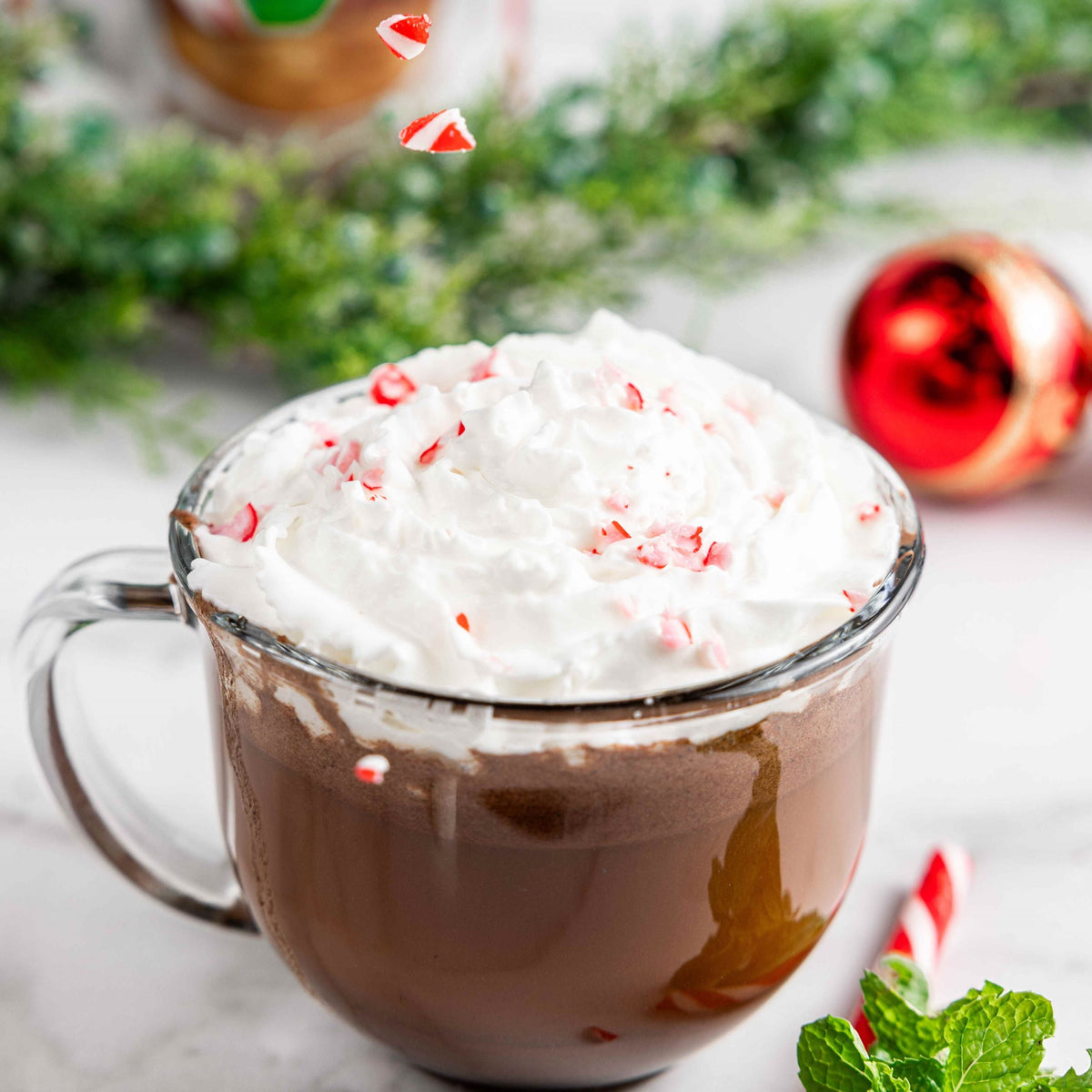 Glass mug of Bare Life peppermint hot cocoa topped with whipped topping and crushed candy canes beside holiday decorations