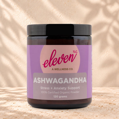 Eleven TLC Organic Ashwagandha Root Powder 120g amber glass jar with a black lid on travertine stone against a warm cream plaster wall, with a soft shadow to the right.