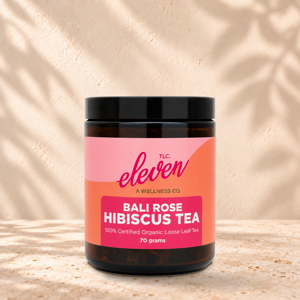 Eleven TLC Bali Rose Loose Leaf Tea | Hibiscus, Ginger & Rose, 70g jar in an amber glass jar with a black lid on a travertine ledge against warm plaster, with a slightly stronger contact shadow beneath the base and a soft shadow to the right.