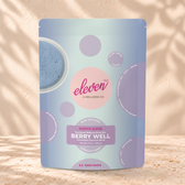 Eleven TLC Berry Well 500g blue and lavender pouch standing on a travertine ledge against warm plaster, with a contact shadow beneath the base and a soft shadow to the right.