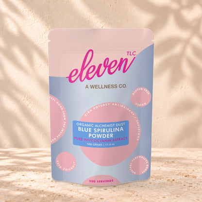 Eleven TLC Organic Blue Spirulina Powder 500g stand-up pouch on travertine stone against a warm cream plaster wall, lit from the left with a soft shadow to the right.