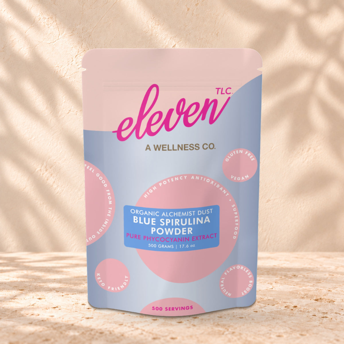 Eleven TLC Organic Blue Spirulina Powder 500g stand-up pouch on travertine stone against a warm cream plaster wall, lit from the left with a soft shadow to the right.