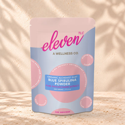 Eleven TLC Organic Blue Spirulina Powder 500g stand-up pouch on travertine stone against a warm cream plaster wall, lit from the left with a soft shadow to the right.
