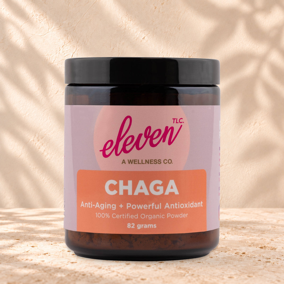 Eleven TLC Organic Chaga Mushroom Powder 82g amber glass jar with a black lid on travertine stone against a warm cream plaster wall, with a soft shadow to the right.
