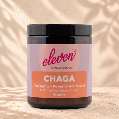 Eleven TLC Organic Chaga Mushroom Powder 82g amber glass jar with a black lid on travertine stone against a warm cream plaster wall, with a soft shadow to the right.