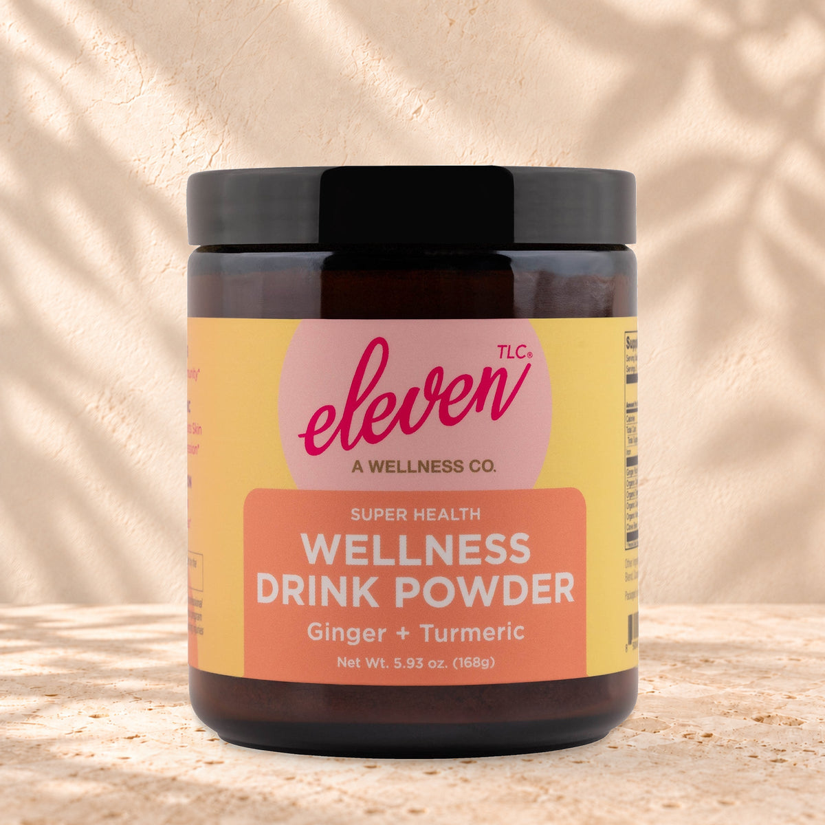 Eleven TLC Organic Ginger Turmeric Golden Latte Mix 168g amber glass jar with a black lid on travertine stone against a warm cream plaster wall, with a soft shadow to the right.
