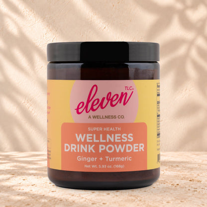 Eleven TLC Organic Ginger Turmeric Golden Latte Mix 168g amber glass jar with a black lid on travertine stone against a warm cream plaster wall, with a soft shadow to the right.
