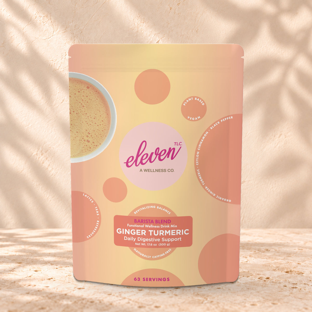 Eleven TLC Organic Ginger Turmeric Golden Milk Latte Base 500g stand-up pouch on travertine stone against a warm cream plaster wall, lit from the left with a soft shadow to the right.