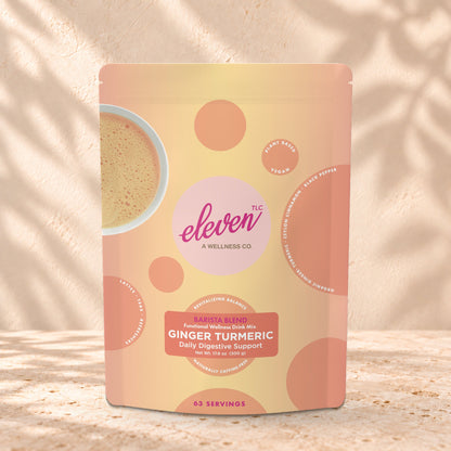 Eleven TLC Organic Ginger Turmeric Golden Milk Latte Base 500g stand-up pouch on travertine stone against a warm cream plaster wall, lit from the left with a soft shadow to the right.