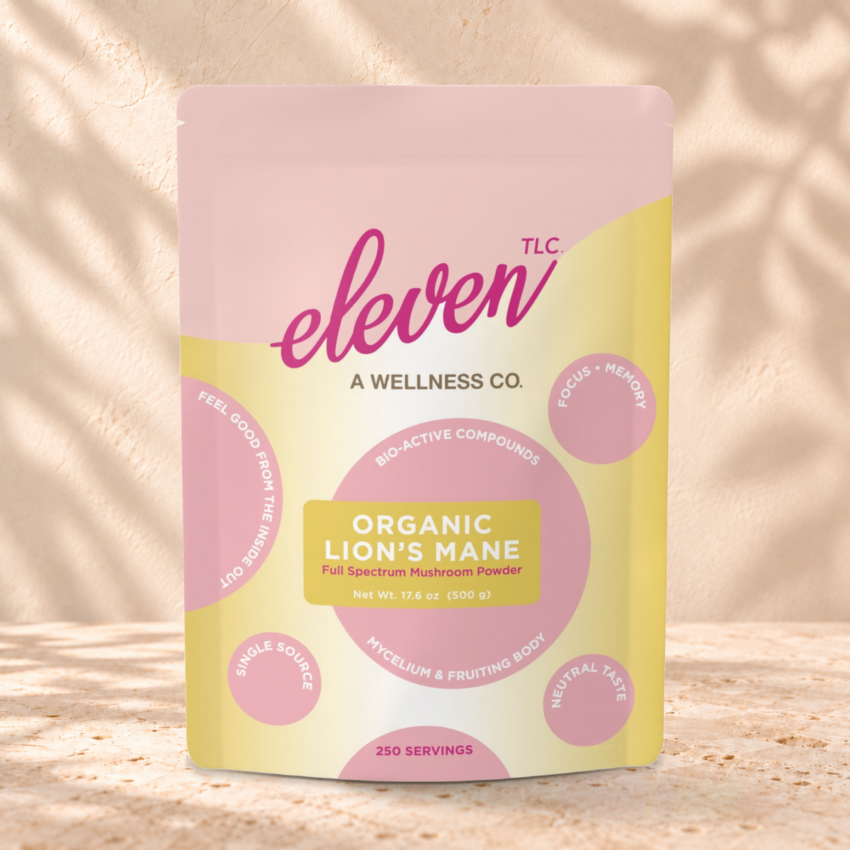 Eleven TLC Lion's Mane 500g yellow and pink pouch standing on a travertine ledge against warm plaster, with a contact shadow beneath the base and a soft shadow to the right.