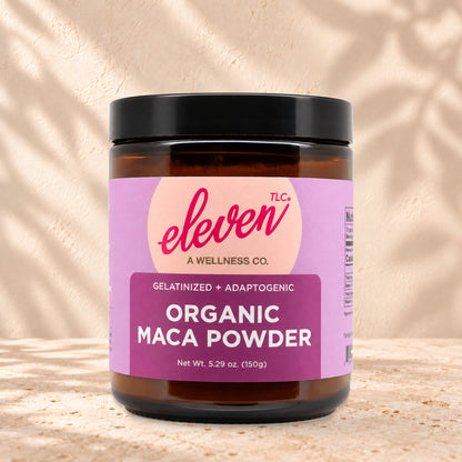 Eleven TLC Organic Gelatinized Maca Root Powder 150g amber glass jar with a black lid on travertine stone against a warm cream plaster wall, with a soft shadow to the right.