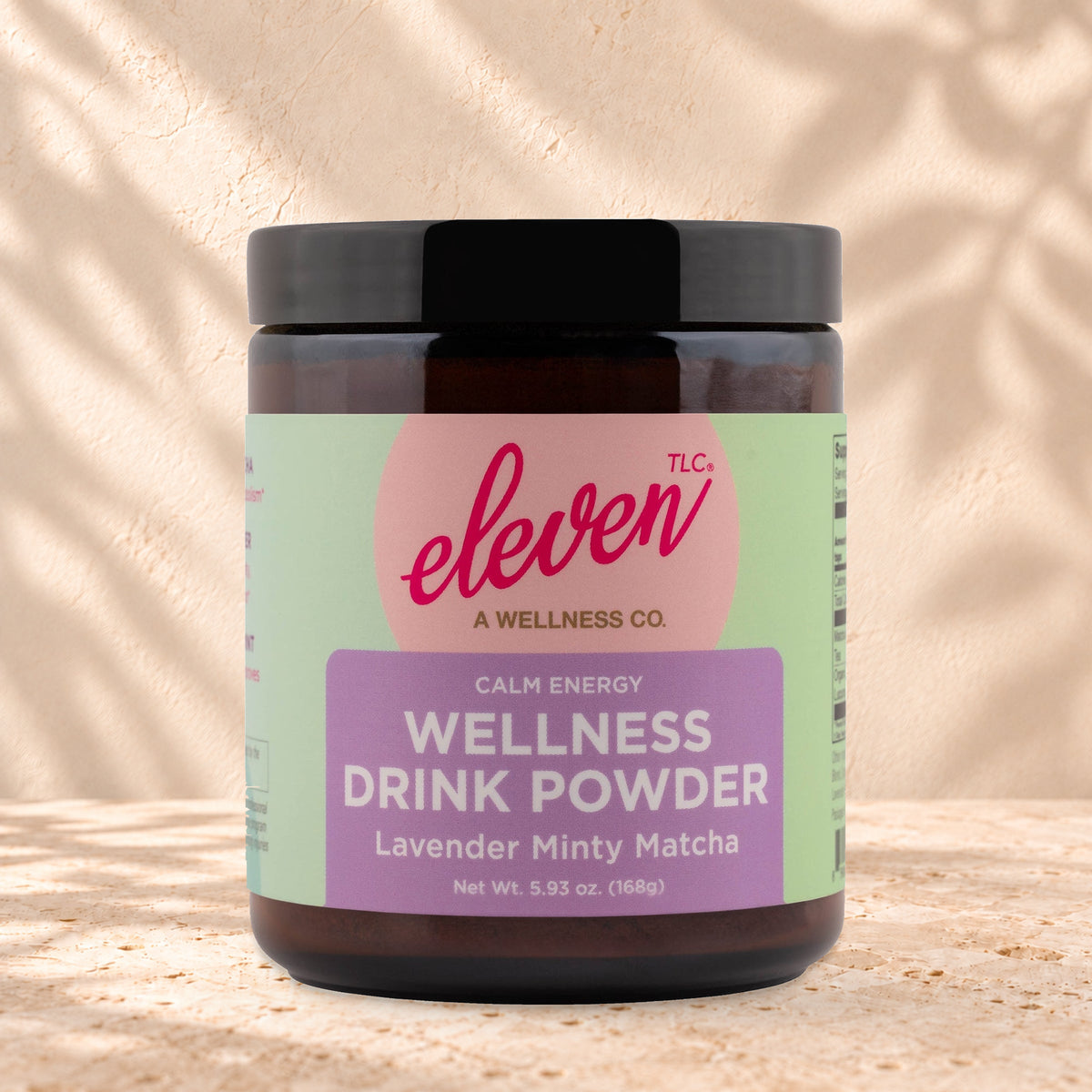 Eleven TLC Minty Lavender Matcha 168g amber glass jar with a black lid on travertine stone against a warm cream plaster wall, with a soft shadow to the right.