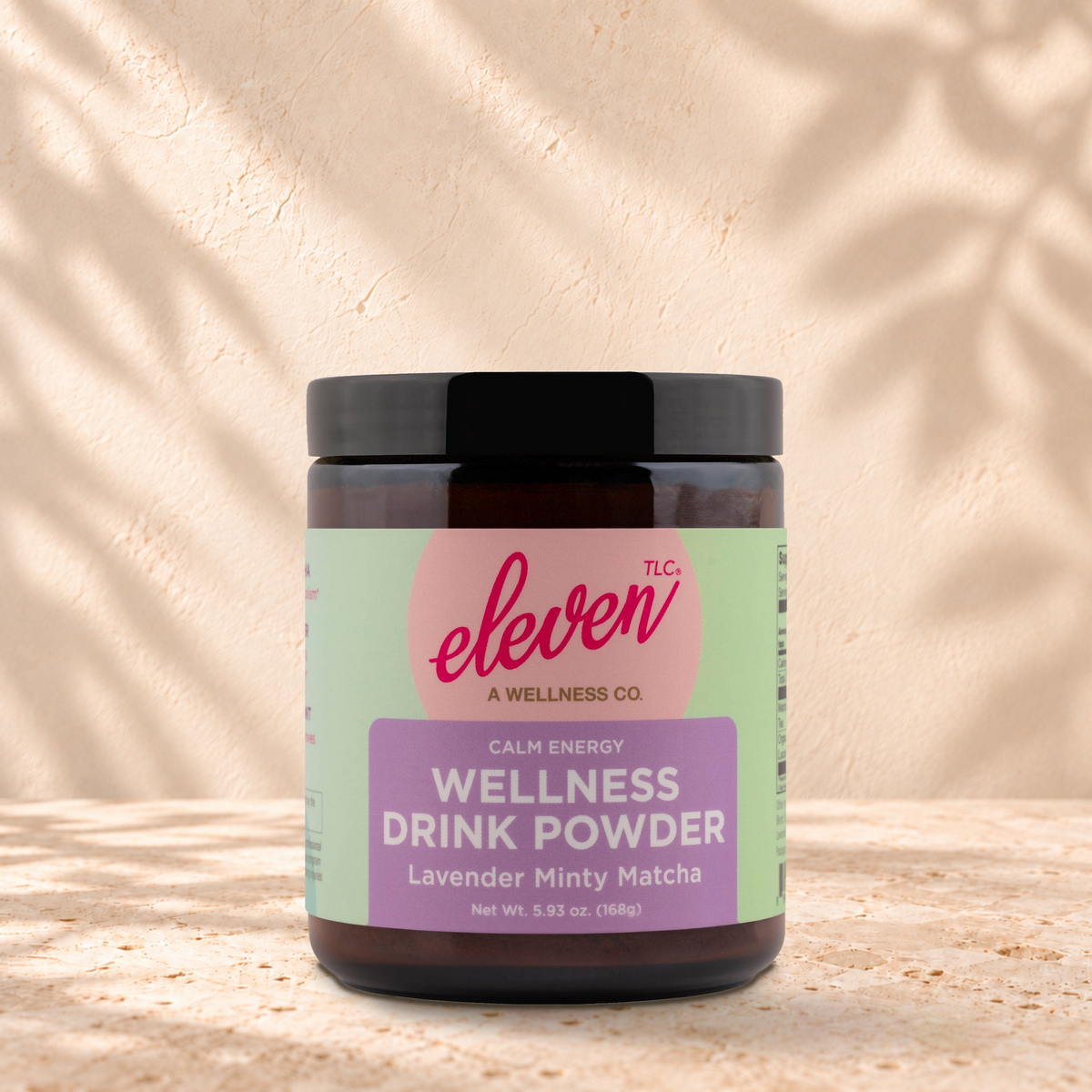 Eleven TLC Minty Lavender Matcha | Calm Focus, 168g jar in an amber glass jar with a black lid on a travertine ledge against warm plaster, with a slightly stronger contact shadow beneath the base and a soft shadow to the right.