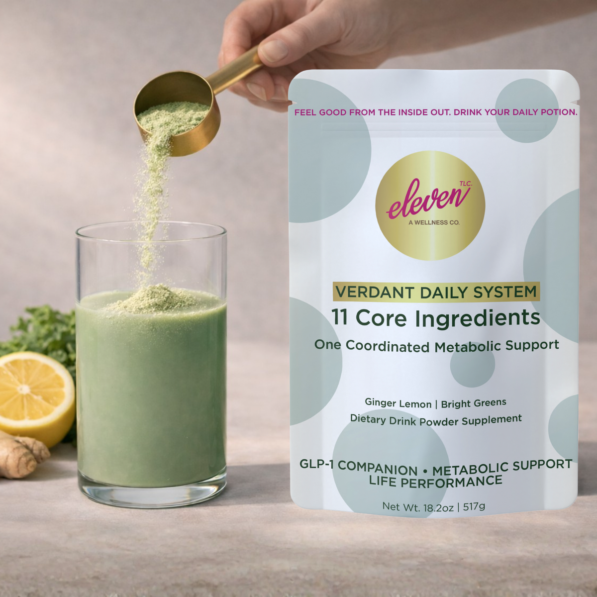 A hand holds a metallic scoop filled with Verdant Daily System powder over a glass of prepared green metabolic beverage. The background features the 517g product pouch, a slice of lemon, and a minimalist, light-toned surface. The lighting is warm and natural, capturing the active preparation phase of the daily signal workflow, showcasing the fine powder consistency and the homogenized liquid blend in a clear glass vessel.