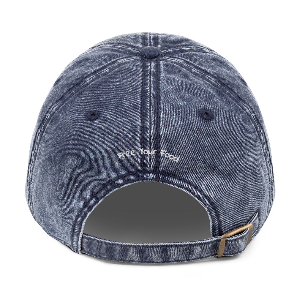 Back view of Bare Life's navy denim Cocoa-Chic Cap with 'Free Your Food' stitched above the adjustable antique brass buckle strap.