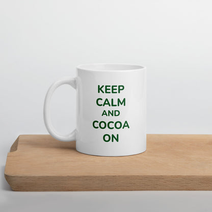 A white ceramic Bare Life mug with the phrase "Keep Calm and Cocoa On" in green lettering sits on a wooden cutting board. The minimalist background emphasizes the sleek design, perfect for cozy hot cocoa moments.
