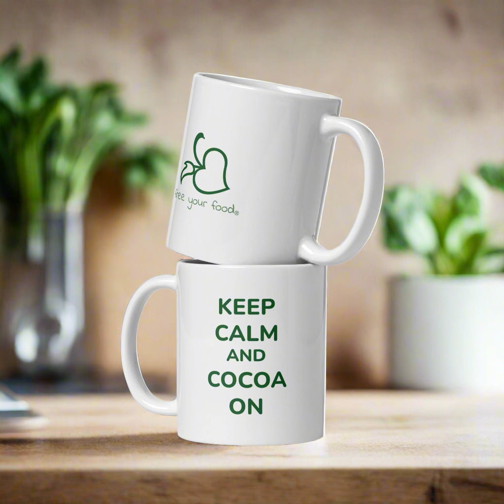 Two white Bare Life mugs stacked on a wooden surface, one featuring the phrase "Keep Calm and Cocoa On" in green text and the other displaying the Bare Life "Free Your Food" logo. A cozy, plant-filled background adds warmth to the scene.