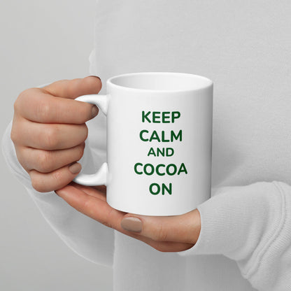 A person wearing a white sweater holds a white Bare Life mug featuring the phrase "Keep Calm and Cocoa On" in green text. The cozy grip and soft background highlight the perfect way to enjoy a warm, dairy-free hot cocoa.