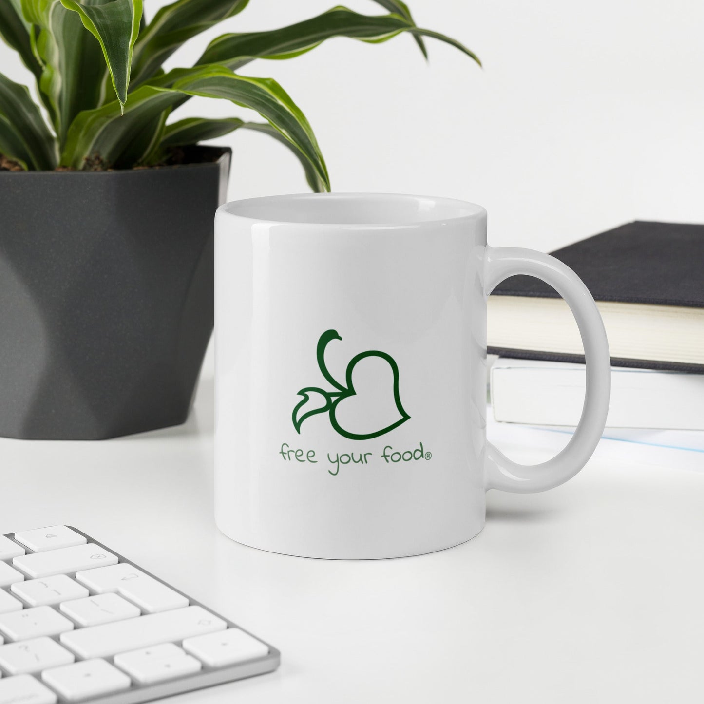 A white Bare Life mug featuring the "Free Your Food" logo sits on a white desk next to a plant, books, and a keyboard. A perfect companion for work breaks, fueling your day with allergen-friendly hot cocoa.